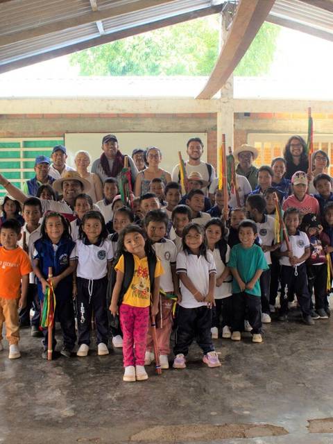 Un grupo grande de niños y adultos posando juntos en un aula con techado de lámina.