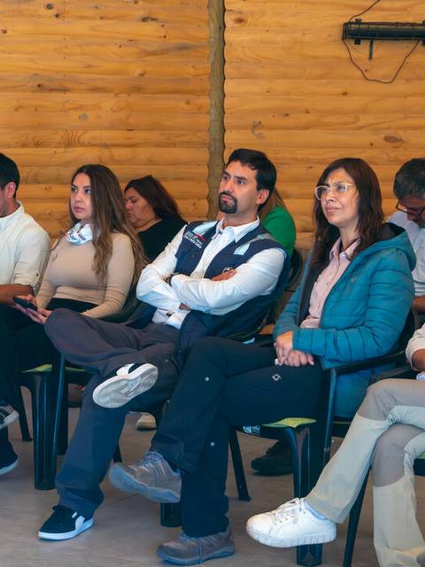 Un grupo de personas sentadas en un salón con paredes de madera, escuchando atentamente.