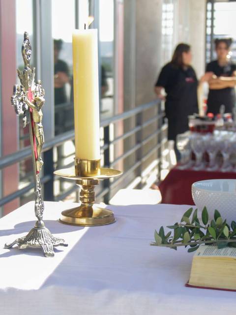 La imagen muestra una mesa dispuesta con un crucifijo, una vela encendida, un cuenco y un libro, en un ambiente preparado para un evento ceremonial.