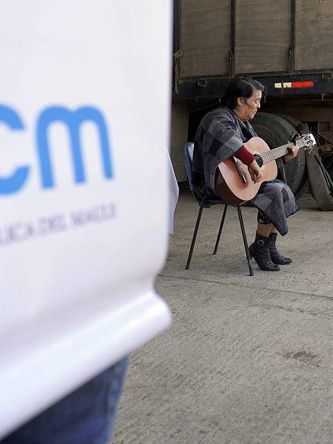 Una mujer toca la guitarra en un lugar al aire libre, mientras se observan mesas con objetos y hay una persona al fondo.