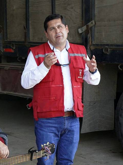 Un hombre de pie con un micrófono habla mientras una mujer sentada toca la guitarra.