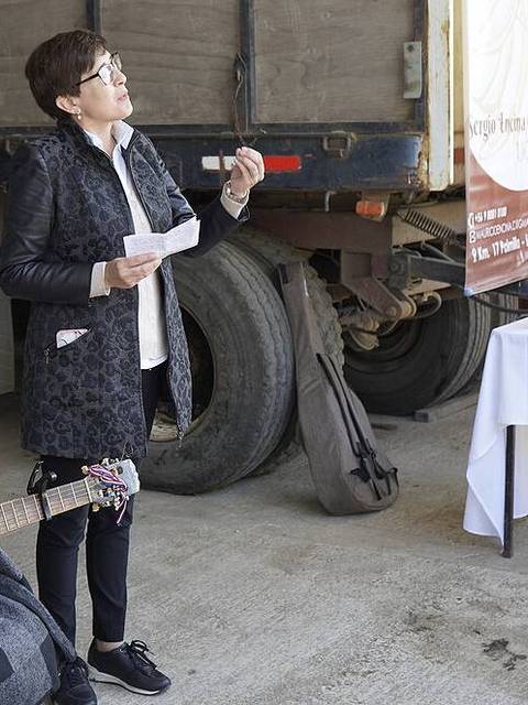 Un grupo de personas se reúne en un evento al aire libre donde una mujer habla mientras otra toca la guitarra.