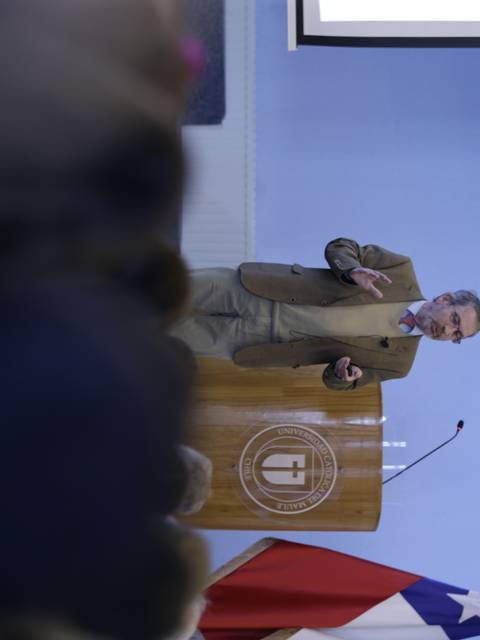 Un conferencista se presenta en un evento mientras una bandera chilena está visible en la parte inferior de la imagen.