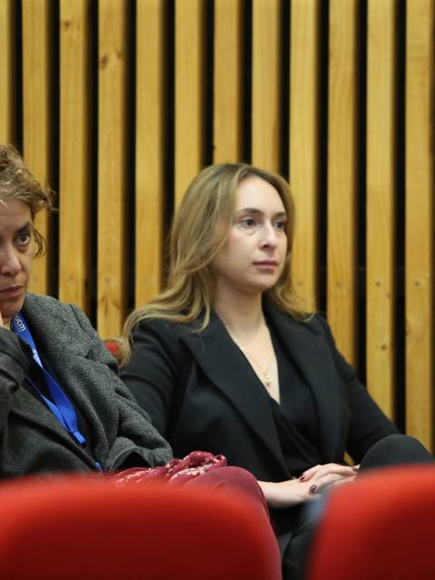 Dos mujeres sentadas en un auditorio con un fondo de paneles de madera.