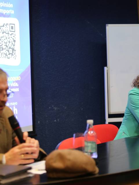 Un hombre habla frente a una audiencia mientras una mujer lo escucha atentamente en un acto público.