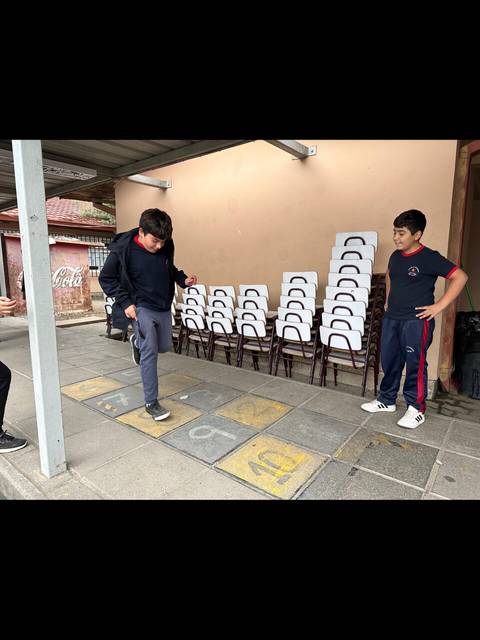 Dos niños juegan en un patio, uno de ellos intentando saltar sobre los cuadros numerados en el suelo.