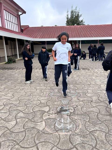 Un grupo de estudiantes juega en el patio escolar mientras una niña salta sobre un dibujo en el suelo.