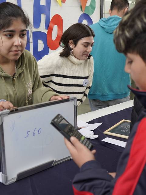 Dos jóvenes interactúan en un evento educativo mientras uno sostiene una pizarra y el otro una calculadora.
