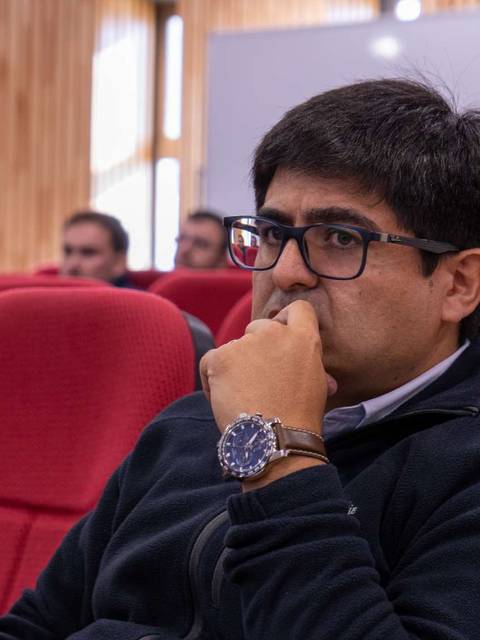 Un hombre se encuentra pensativo en un aula con asientos rojos y una pizarra al fondo.