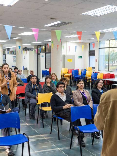 Una persona habla frente a un grupo de asistentes en un evento en un aula decorada con banderines de colores.