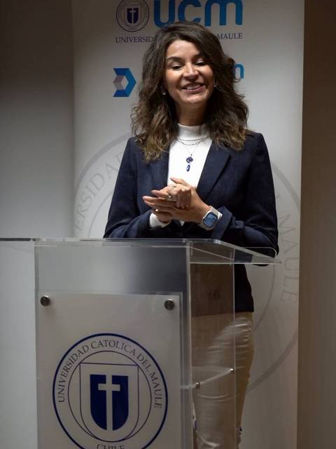 Una mujer sonriente se encuentra detrás de un atril, hablando en un evento académico.