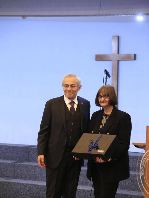Dos personas posan juntas en un evento formal frente a un fondo con una cruz.