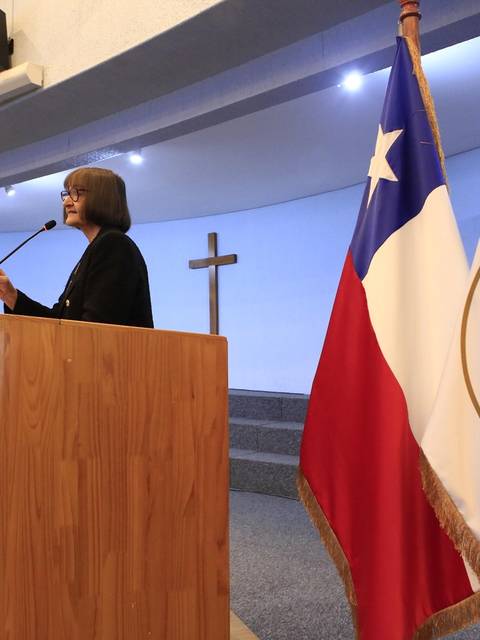 Una mujer habla desde un podio en un evento en la Universidad Católica de Chile, con una bandera chilena y el logo de la universidad en el fondo.