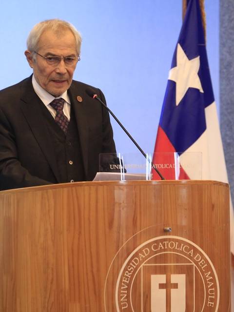 Un hombre mayor está dando un discurso en un podio con banderas de Chile y la universidad al fondo.