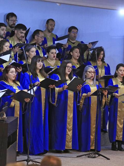 Un coro de personas vestido con túnicas azules y doradas se presenta frente a un piano y una directora.