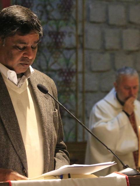 Un hombre lee en un acto religioso mientras un sacerdote lo observa al fondo.