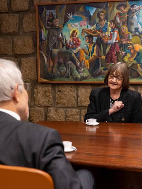 Una conversación entre dos personas en una sala con un mural colorido al fondo.