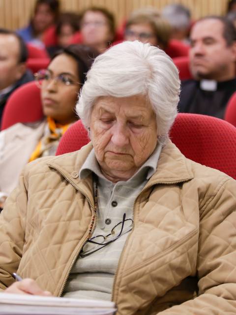 Un grupo de personas sentadas en un auditorio, con una mujer mayor concentrada en tomar notas en primer plano.