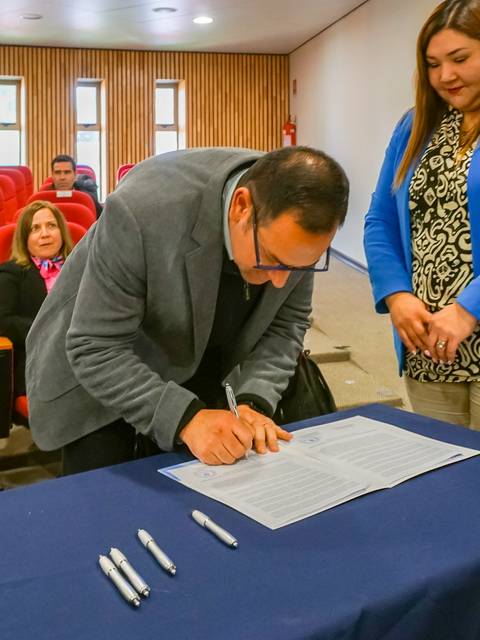 Un hombre firma un documento en un evento, mientras una mujer observa y un grupo de personas asiste al fondo.