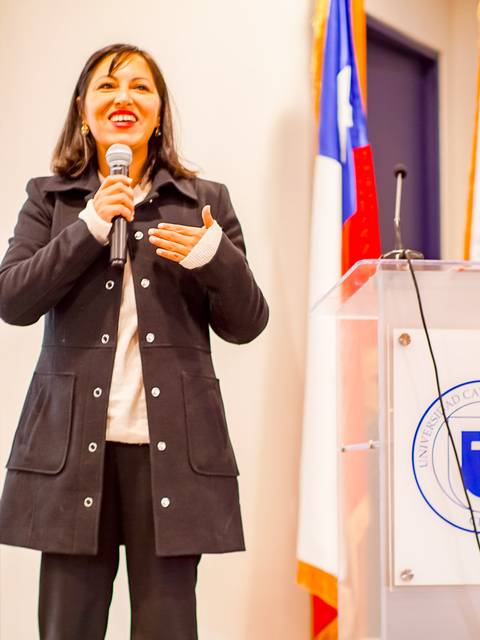 Una mujer de pie habla con entusiasmo frente a un público en un evento.