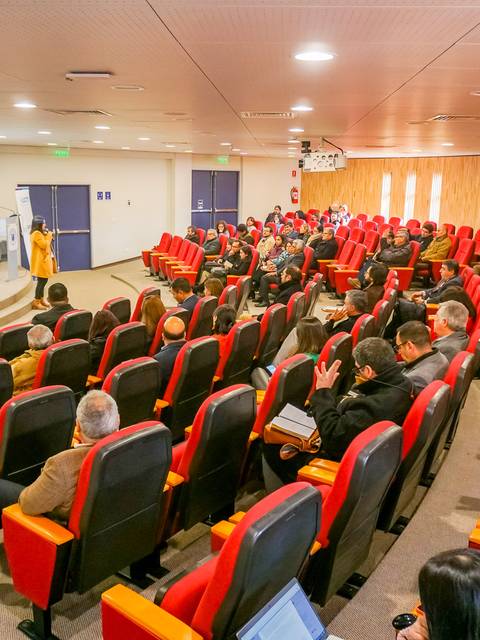 Una sala de conferencias con asientos rojos y un público asistiendo a una presentación.