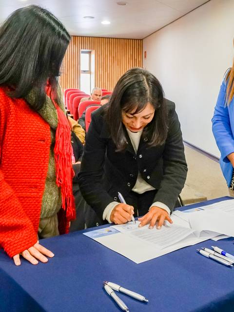 Tres mujeres en un acto formal, donde una de ellas está firmando unos documentos en una mesa.