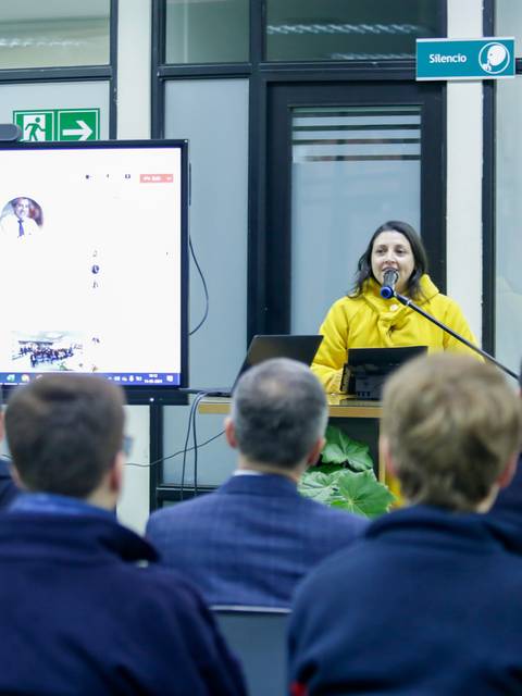 Una presentadora habla frente a un grupo de personas en una sala de conferencias.