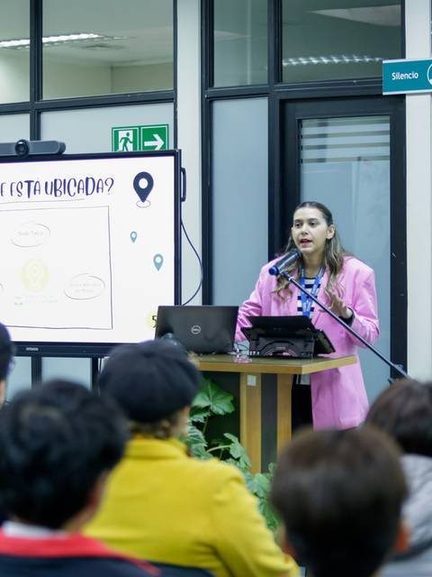Una mujer está dando una presentación frente a un público en un salón con pantallas y un proyector.