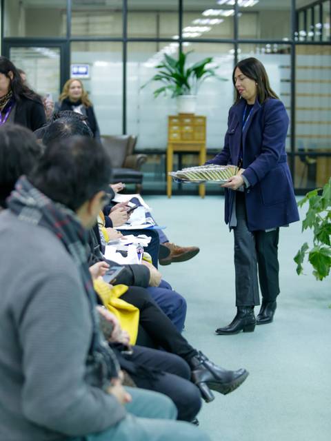 Una mujer presenta un objeto en una sala con personas sentadas y plantas de fondo.