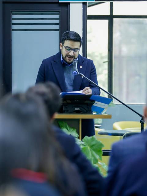 Una persona está leyendo en un micrófono frente a un público en un entorno académico.