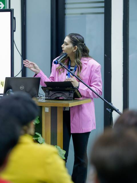 Una mujer con un blazer rosa está presentando en un escenario mientras usa un micrófono.