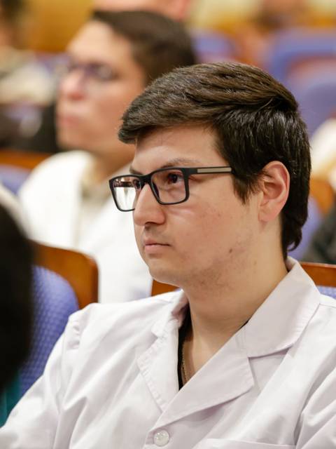 Una persona con bata blanca y gafas escucha atentamente en un auditorio lleno de asistentes.