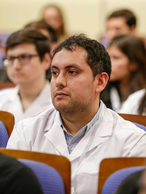 Un grupo de personas en una conferencia, destacando a un hombre con bata blanca que escucha atentamente.