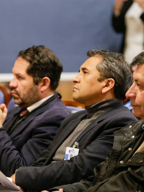 Tres hombres están sentados, escuchando atentamente en un evento.