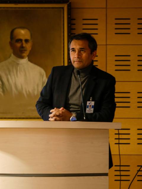 Un hombre de pie frente a un podio, con un retrato enmarcado de un individuo detrás de él.