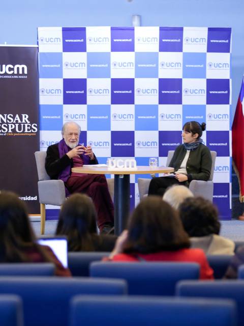 Un evento académico con un panel de discusión en el que participan dos personas, rodeados de una audiencia en un auditorio.