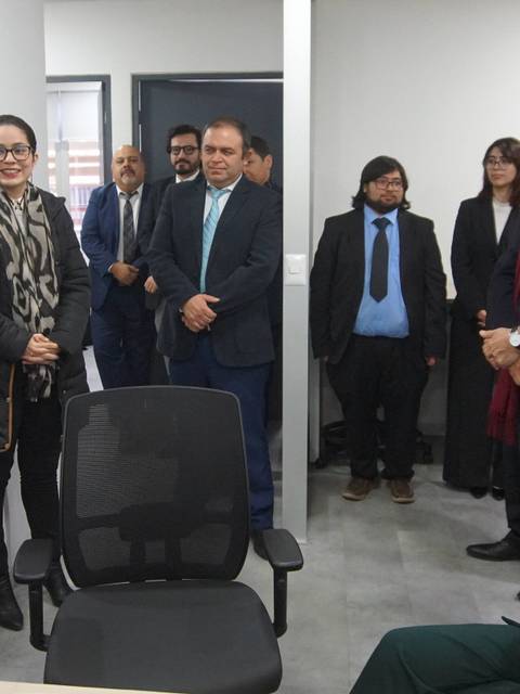 Un grupo de personas vestidas formalmente se reúnen en una oficina moderna, escuchando a un orador que se encuentra de pie frente a ellas.
