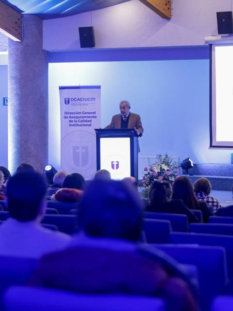 Un orador está dando un discurso en un evento institucional ante una audiencia sentada.