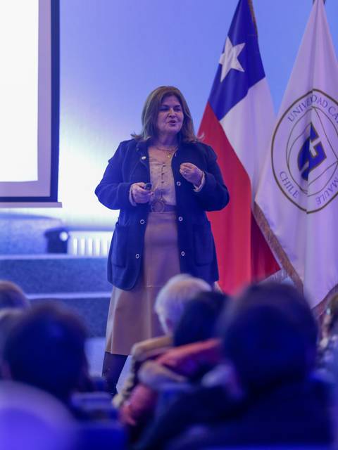 Una mujer de pie presenta en un escenario con banderas detrás en un evento institucional.