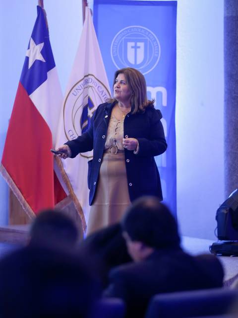 Una mujer presenta en un escenario mientras se observa una bandera chilena y un estandarte institucional en el fondo.