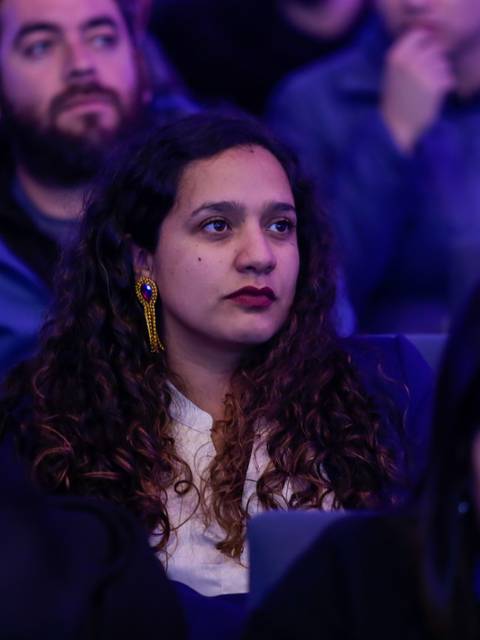Una mujer con cabello rizado y aretes se encuentra sentada entre un grupo de personas en un evento.