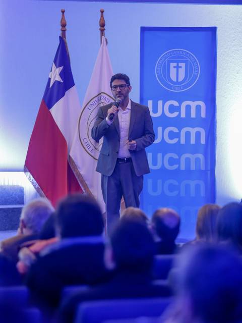 Un hombre de pie habla en un evento con banderas chilena y de la universidad detrás.