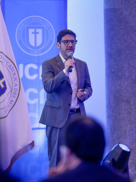 Un hombre vestido de traje habla con un micrófono en un evento, mientras hay una bandera de fondo.