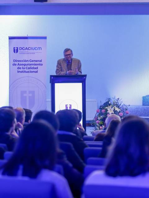 Un conferencista está hablando en un evento frente a una audiencia en un salón decorado adecuadamente.