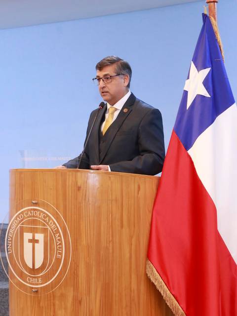 Un hombre de pie frente a un podio con banderas de Chile y un símbolo de una universidad, durante un evento formal.