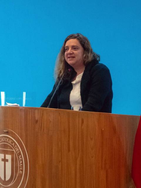 Una mujer está hablando en un podio frente a una bandera chilena y una cruz en una pared azul.