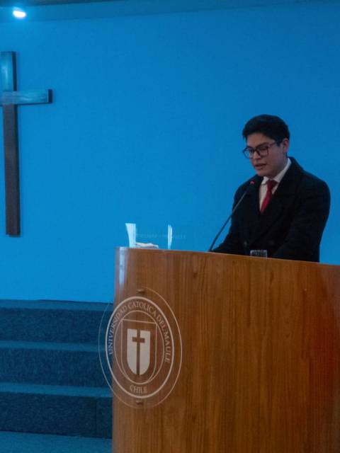 Un hombre presenta en una conferencia en un auditorio con banderas y un altar detrás.