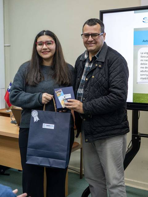Una mujer y un hombre se encuentran en un evento, donde la mujer recibe un reconocimiento y sostiene un regalo.