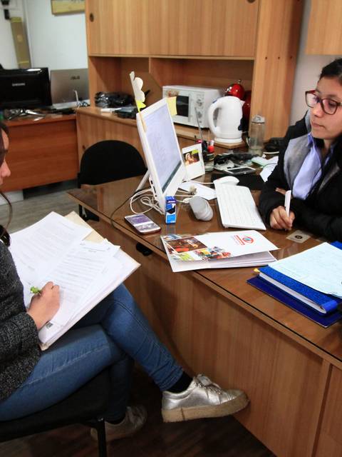 Dos personas conversan en una oficina mientras revisan documentos y utilizan computadoras.