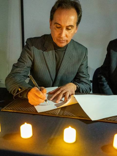 Un hombre vestido con un traje oscuro firma un documento frente a unas velas encendidas.